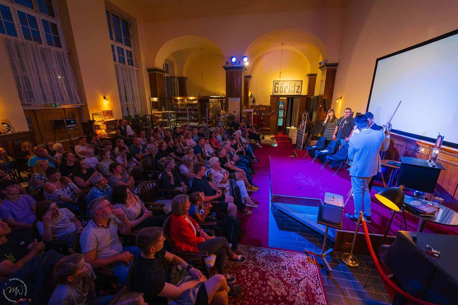 Weitwinkelaufnahme der Bühne mit Thomas, Vincent und Publikum.