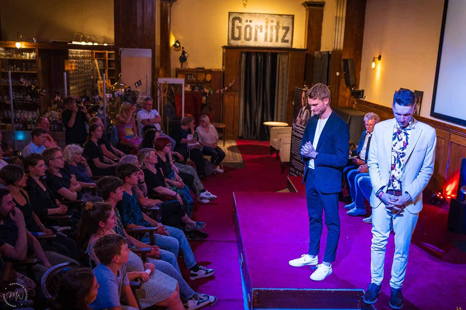 Thomas und Vincent stehen auf der Bühne, die Zuschauer sind im Hintergrund zu sehen.