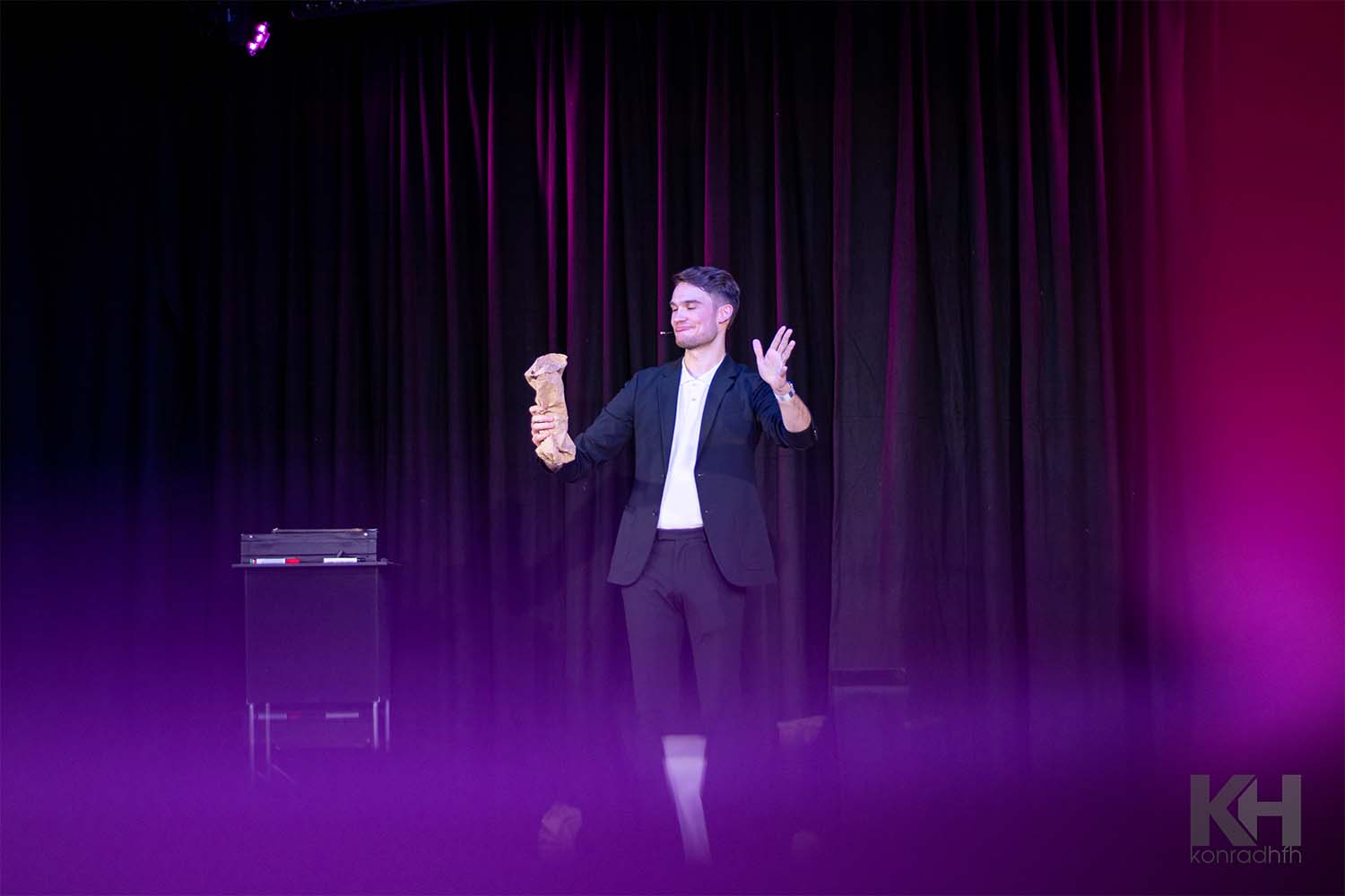 Vincent Frommer steht mit verschmitztem Blick auf der Bühne und hält eine Papiertüte in der Hand