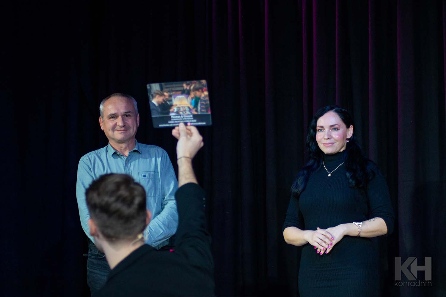 Zwei Zuschauer auf der Bühne betrachten eine hochgehaltene Karte von Vincent Frommer