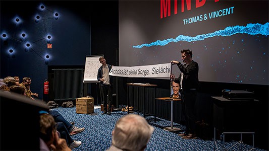 Thomas Majka und Vincent Frommer rollen gemeinsam eine Papierrolle auseinander, auf der eine Vorhersage steht, während der Mindtrix-Show in Bautzen