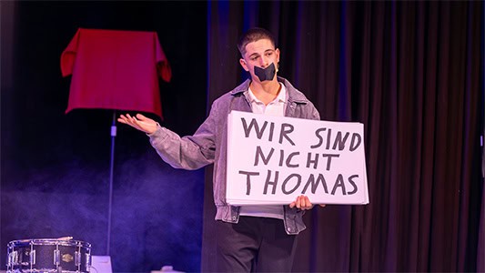Benito hält ein Schild mit der Aufschrift wir sind nicht Thomas, hat den Mund zugeklebt und steht mit Merlin auf der Bühne im Carte Blanche Petit Theater Dresden