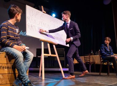 Vincent Frommer auf der Bühne vor einer großen Tafel mit zwei Zuschauern während einer interaktiven Mentalmagie-Show