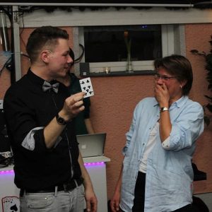 Thomas Majka hält eine Spielkarte hoch, während eine überraschte Zuschauerin daneben mit offenem Mund staunt