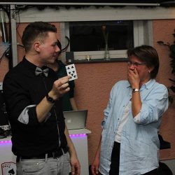 Thomas Majka hält eine Spielkarte hoch, während eine überraschte Zuschauerin daneben mit offenem Mund staunt